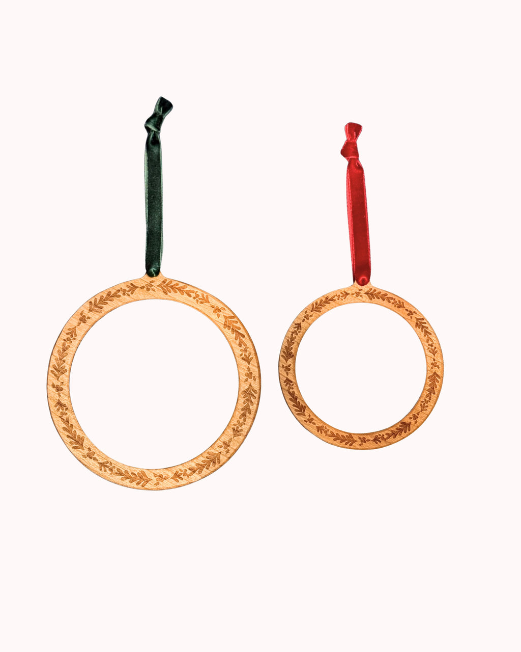Two circular wooden self finishing Christmas ornaments with engraved holly leaf patterns, one larger with a dark green hunter velver hanging ribbon and one smaller with a red velvet hanging ribbon, shown on a plain white background.