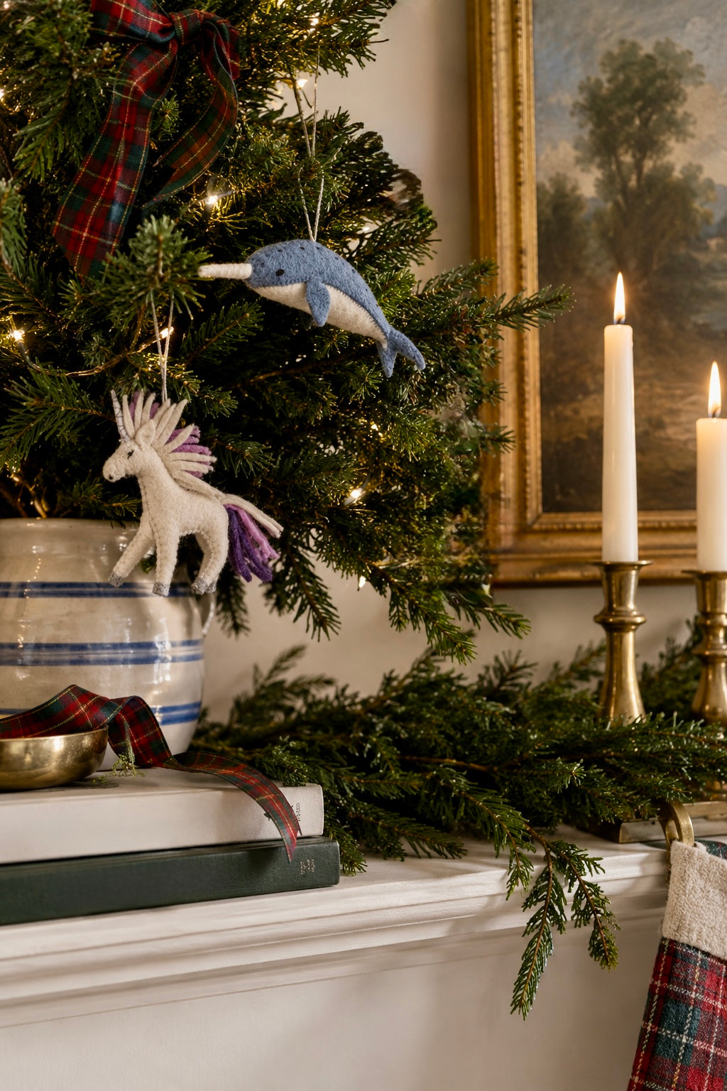 A cozy holiday mantel scene with a green felt unicorn ornament hanging on a decorated Christmas tree, alongside a blue felt whale ornament, a striped ceramic pot, lit white taper candles in brass holders, and a framed landscape painting in the background.