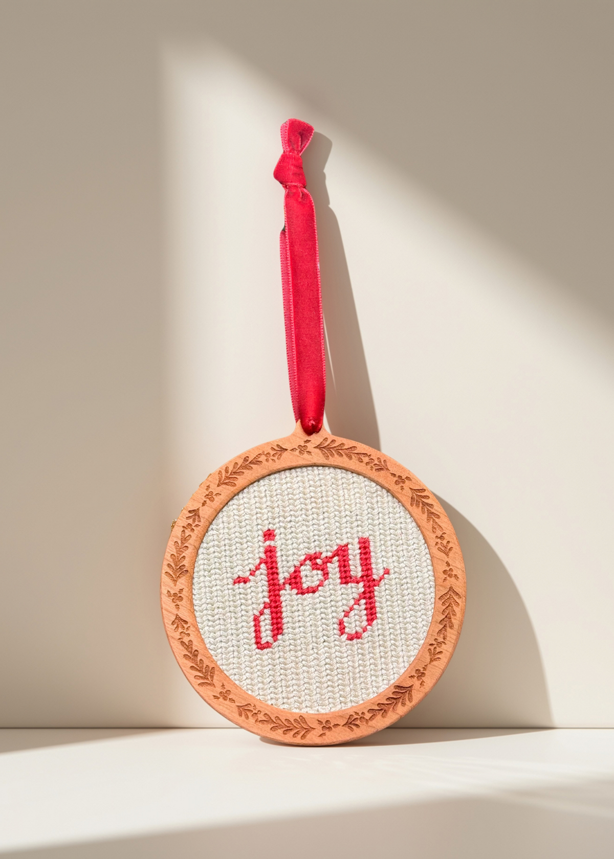 A round needlepointed hanging ornament with the word "joy" stitched in red on a white fabric background, framed by an engraved holly design on our wooden self finishing ornaments hanging from a velvet red ribbon, shown against a warm off-white wall with strong sunlight shadows.